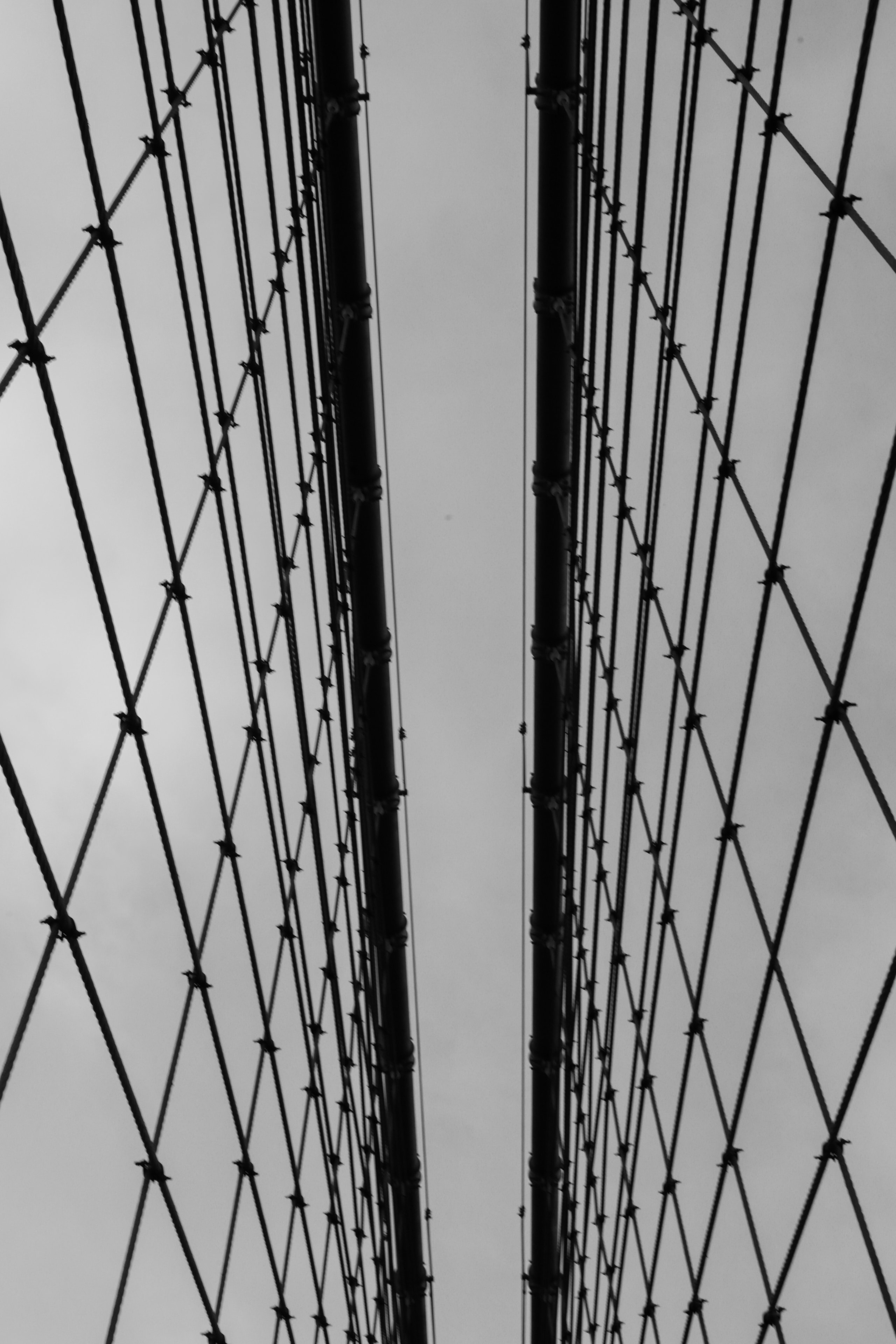 Close-up abstract view looking up at Brooklyn Bridge twin towers with geometric cable web pattern against grey sky