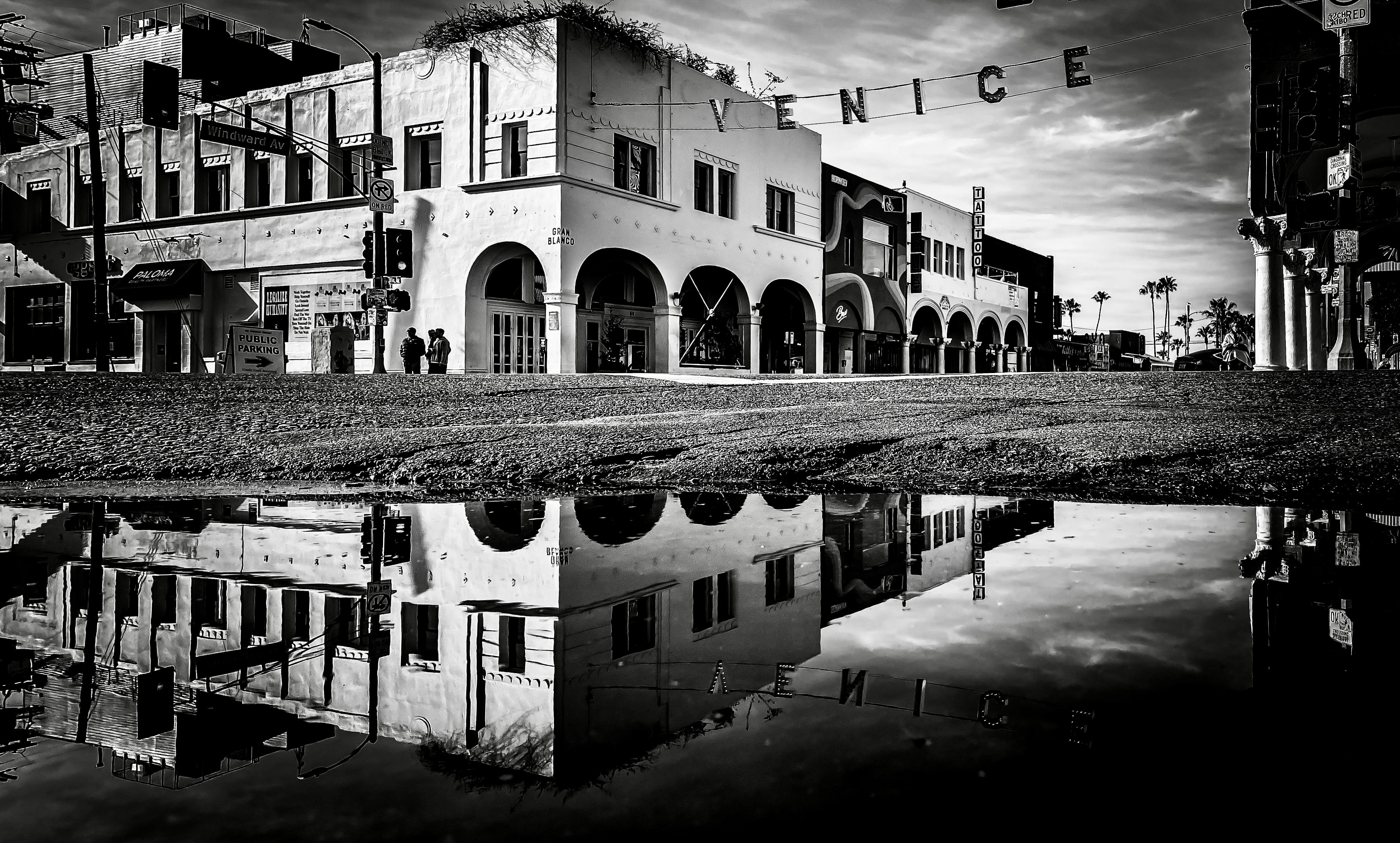 Venice Beach historic arched storefronts with VENICE sign perfectly mirrored in a rain puddle on the street below under dramatic cloudy sky