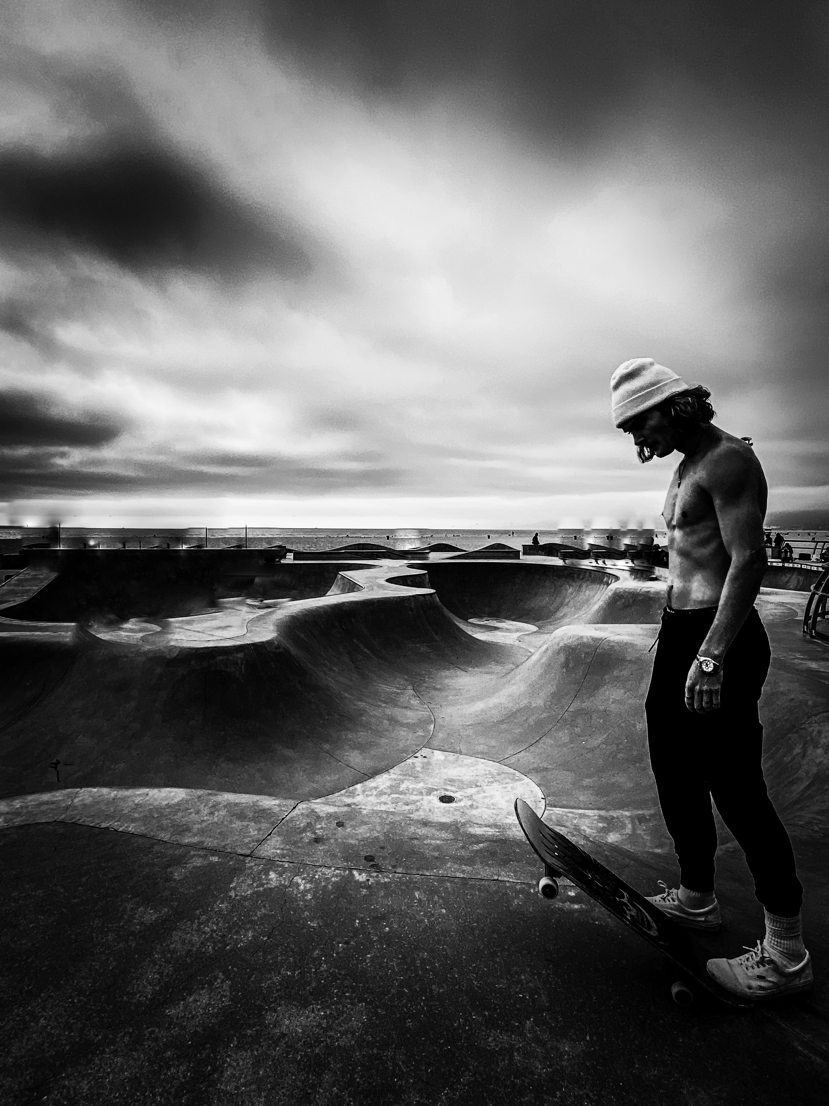 Shirtless skateboarder standing at Venice Beach skate park under dramatic stormy sky and ocean backdrop