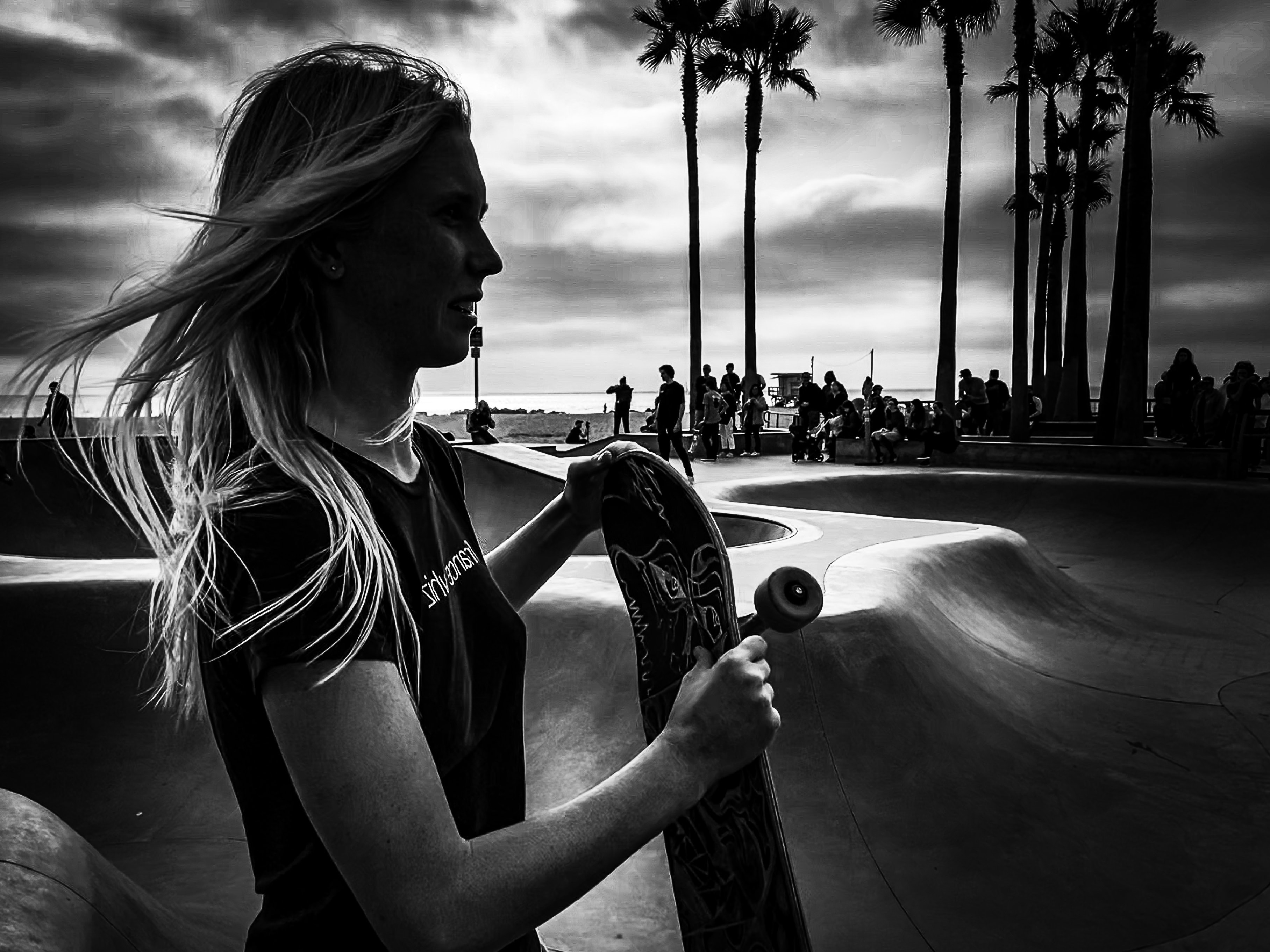 Young woman holding skateboard at Venice Beach skate park with palm trees and crowd silhouetted behind