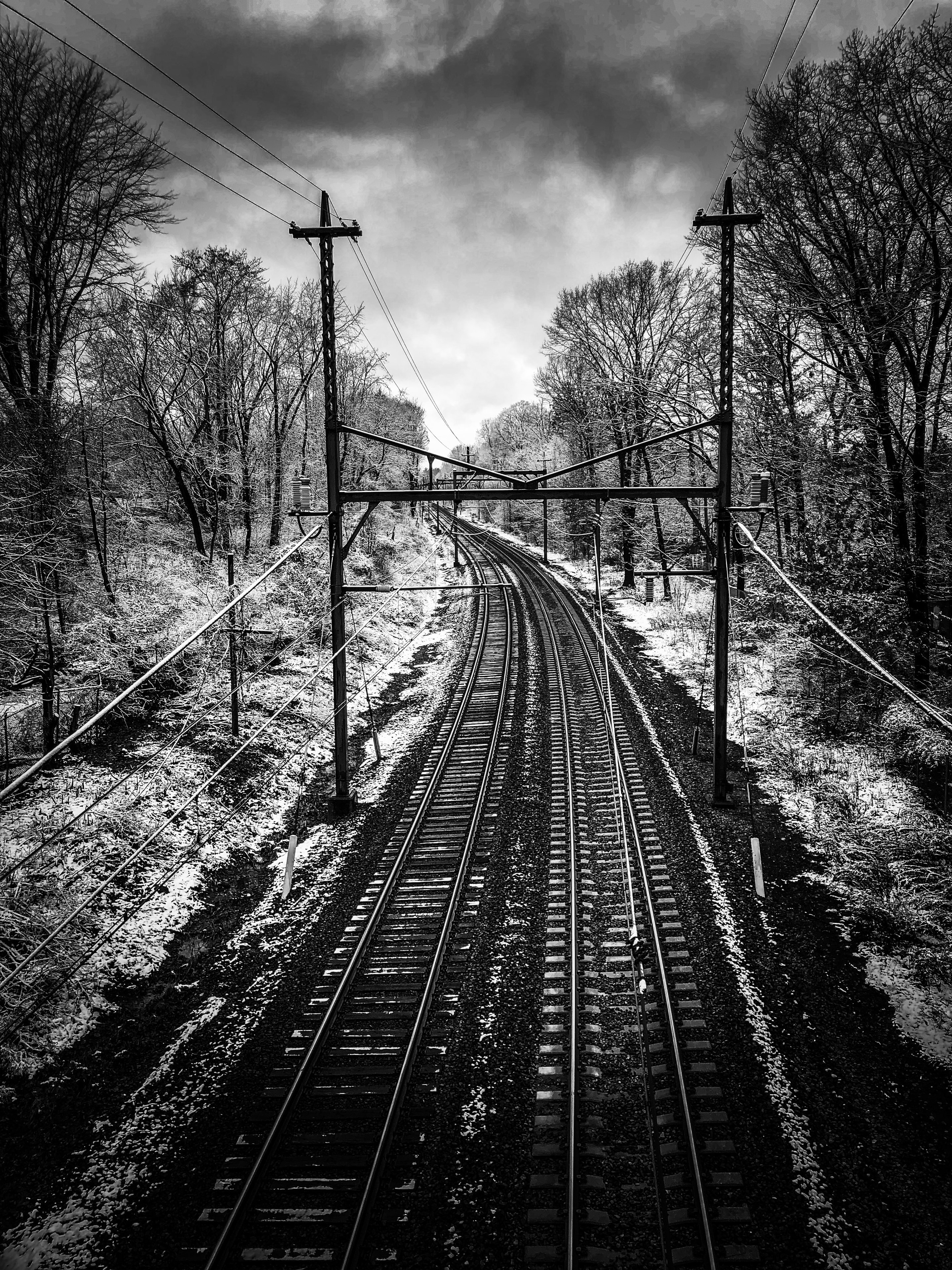 Railroad tracks curving through snow-dusted bare trees under dramatic stormy sky with utility poles