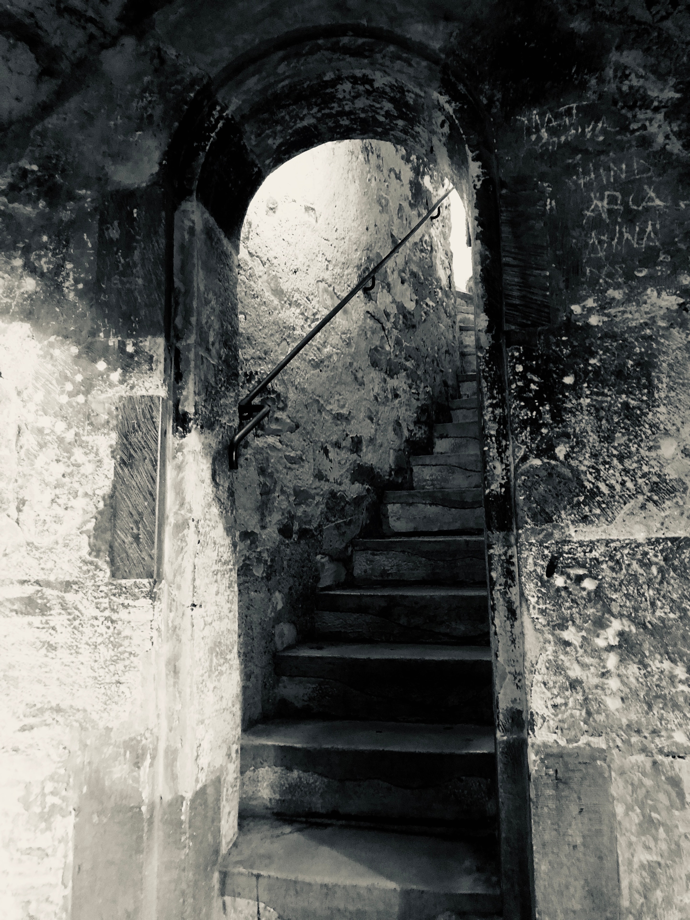 Ancient weathered stone archway framing a dark staircase with iron handrail and crumbling walls