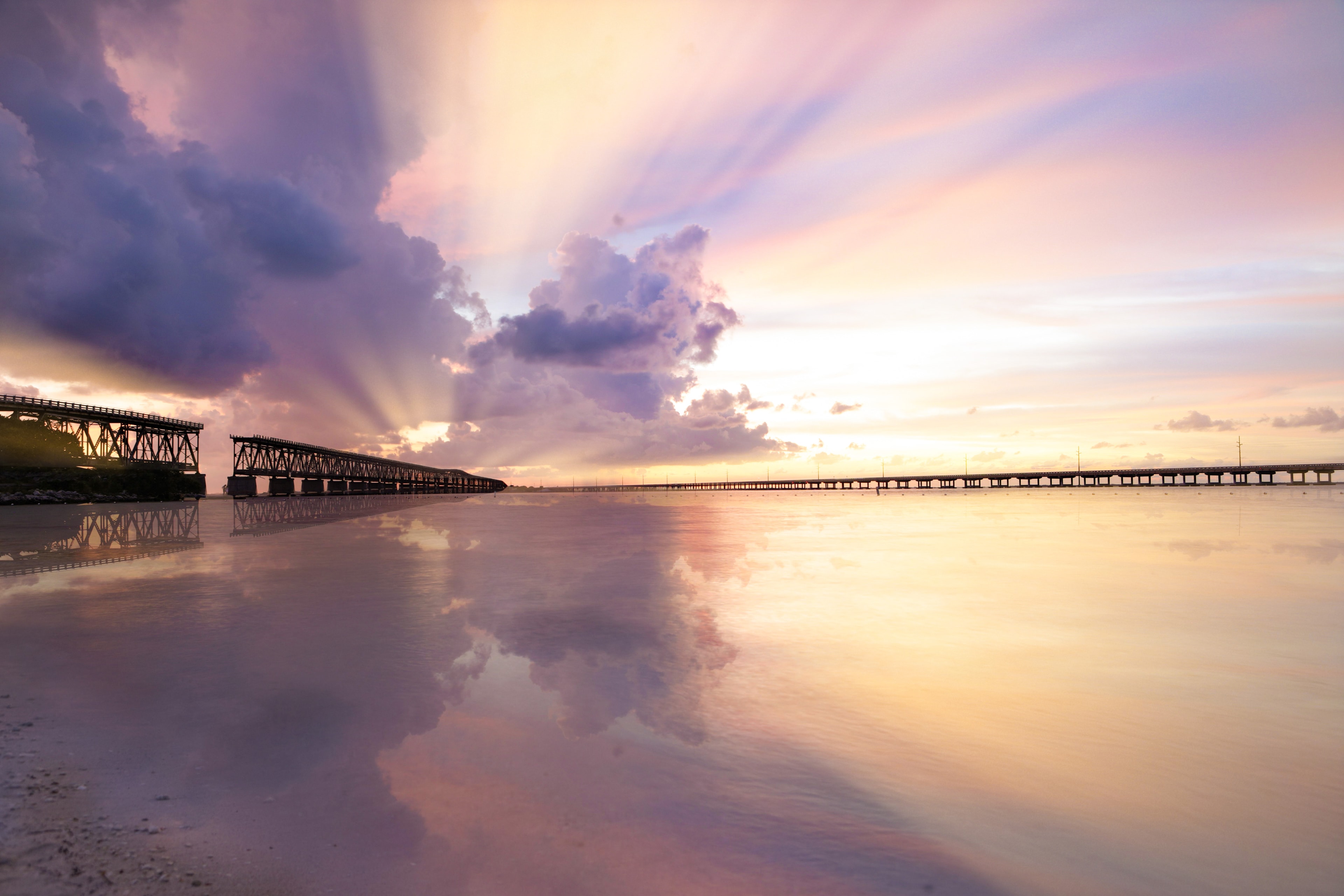 Long pier and bridge spanning calm water at sunset with dramatic purple and golden clouds