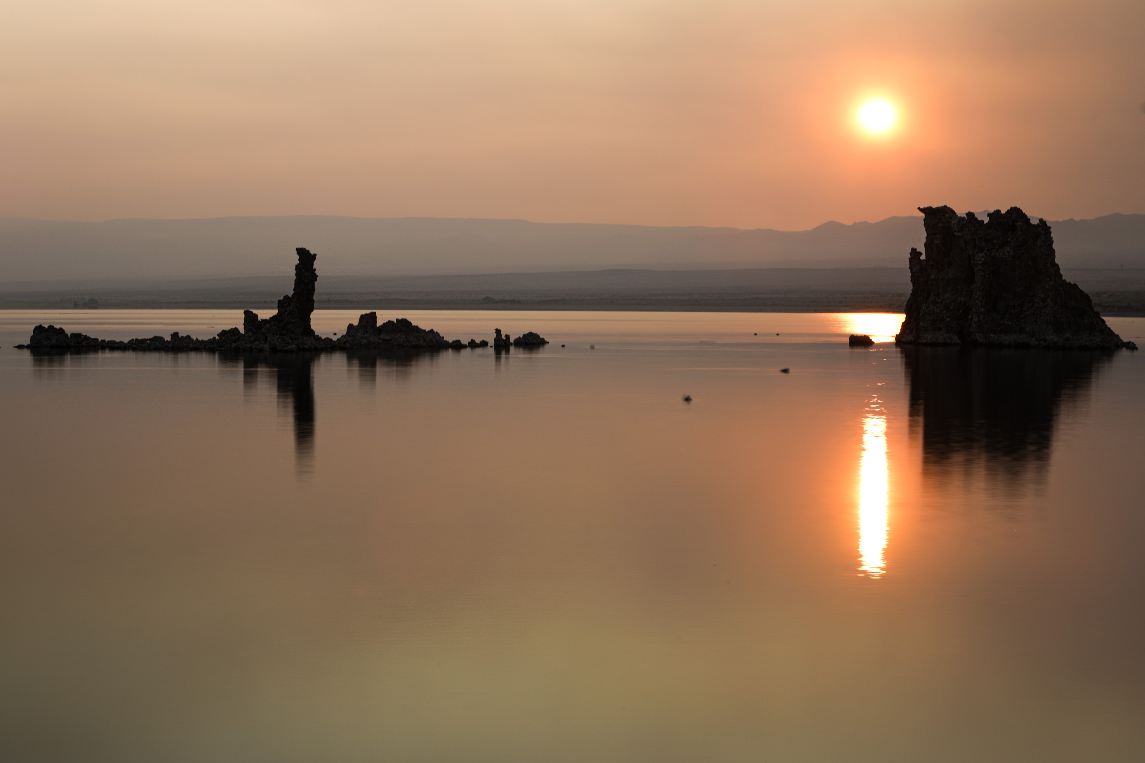 Tufa rock formations silhouetted against hazy orange sunset reflected in still lake