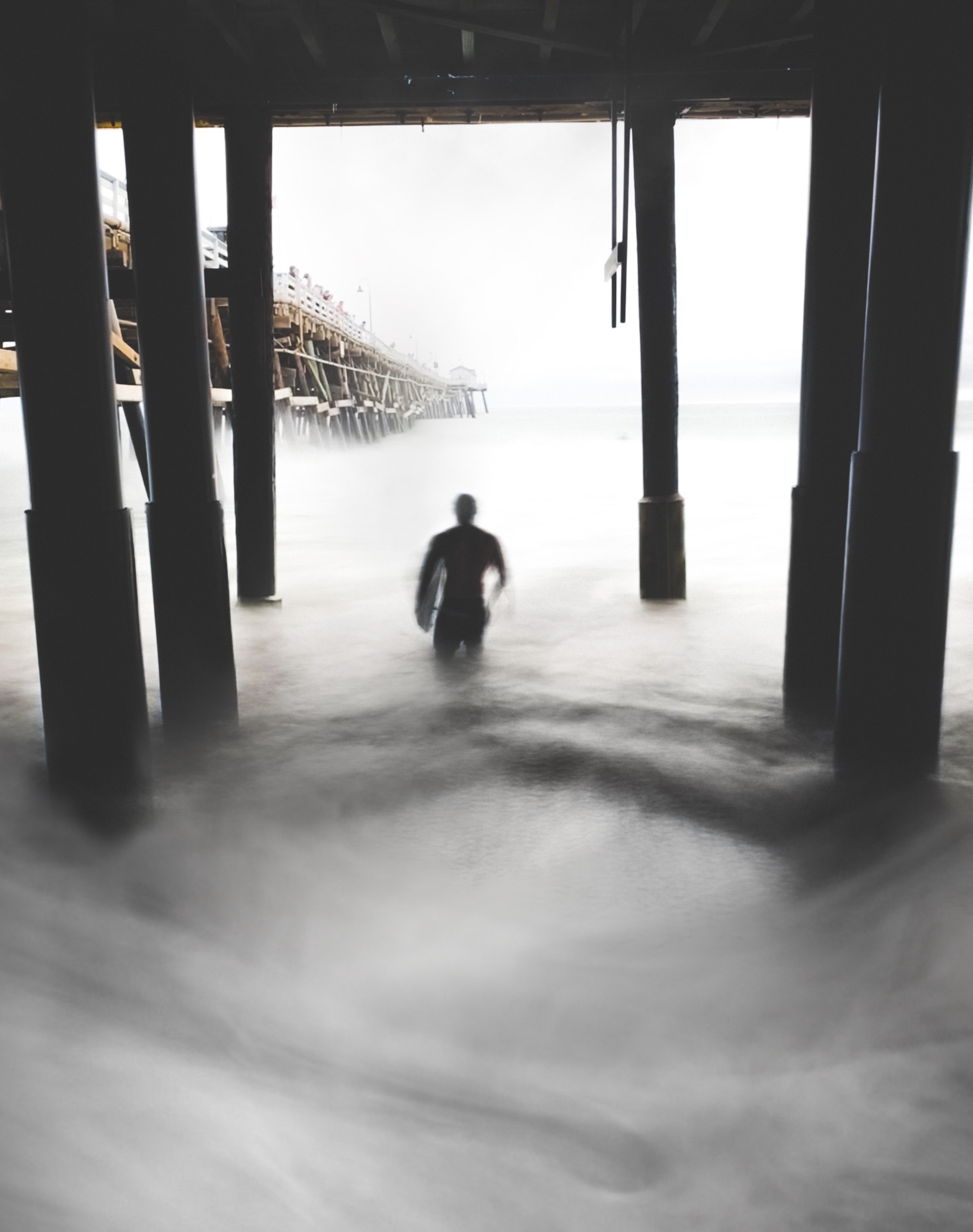 Surfer silhouette wading through swirling ocean mist beneath wooden pier pillars long exposure