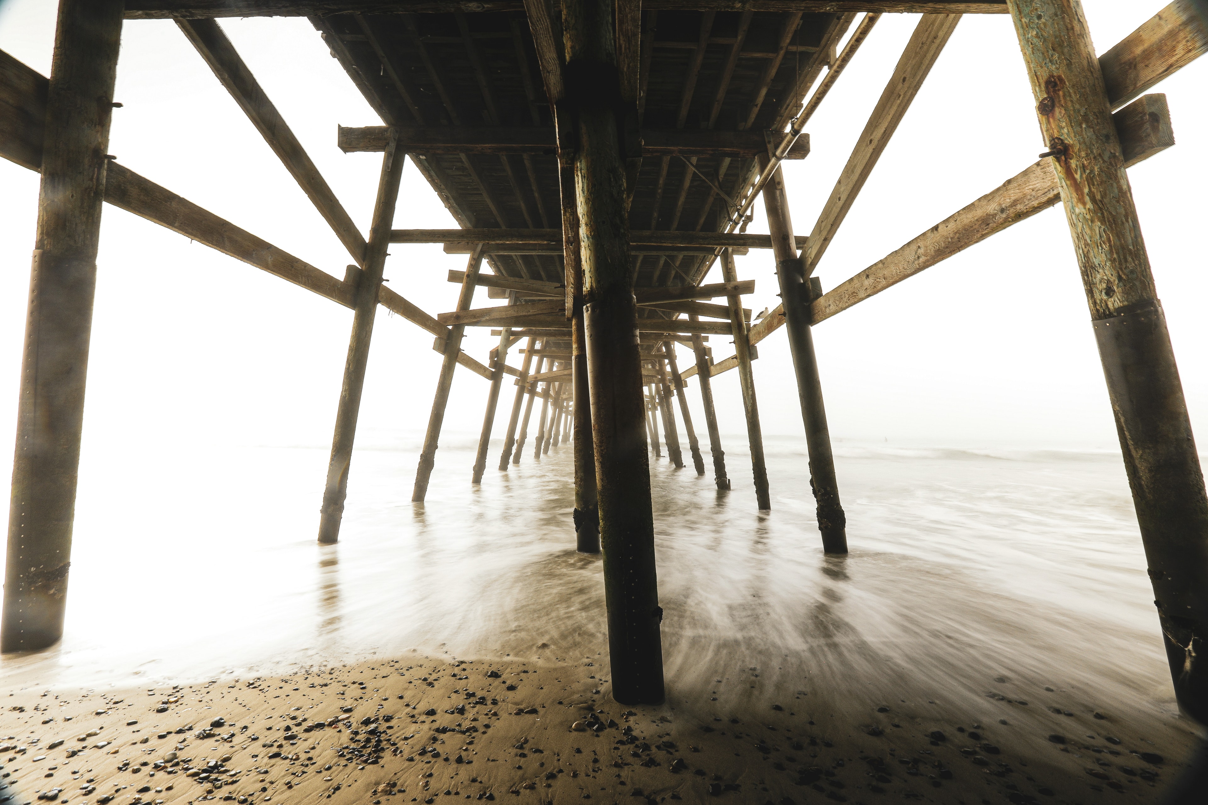 Looking under a weathered wooden pier with silky long-exposure waves washing around the pilings receding into misty light