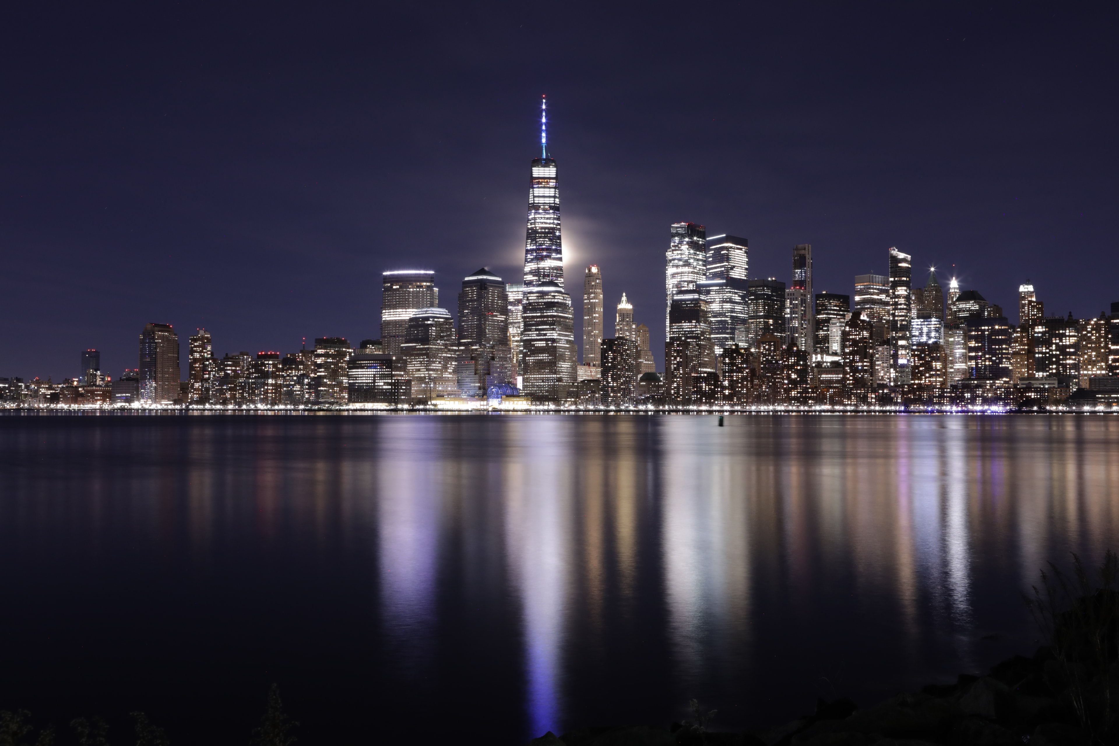 Lower Manhattan skyline at night with full moon rising behind One World Trade Center and colorful light reflections on the Hudson River