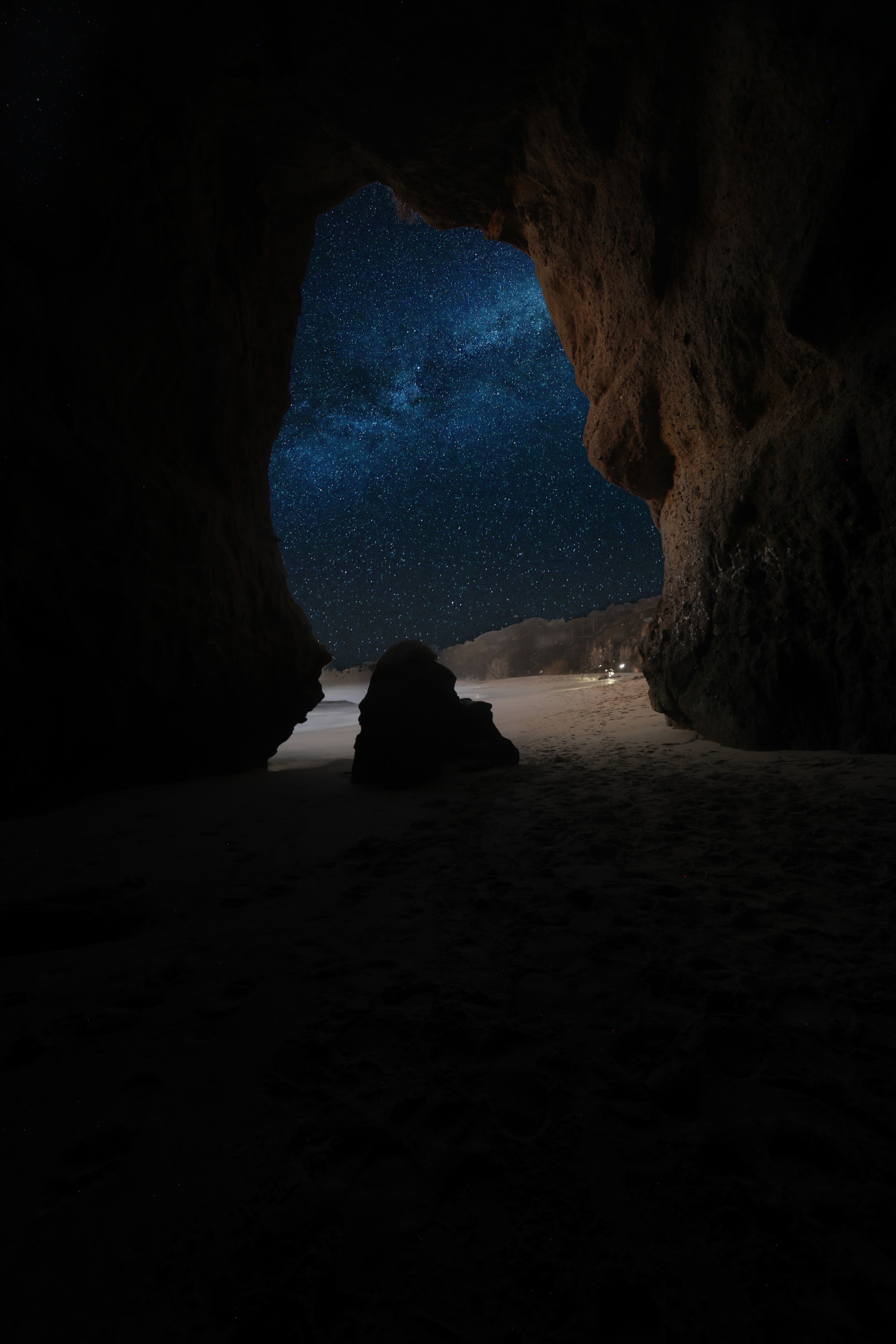 View from inside a dark sea cave looking out through a jagged rock opening to a starry night sky with the Milky Way visible above a sandy beach