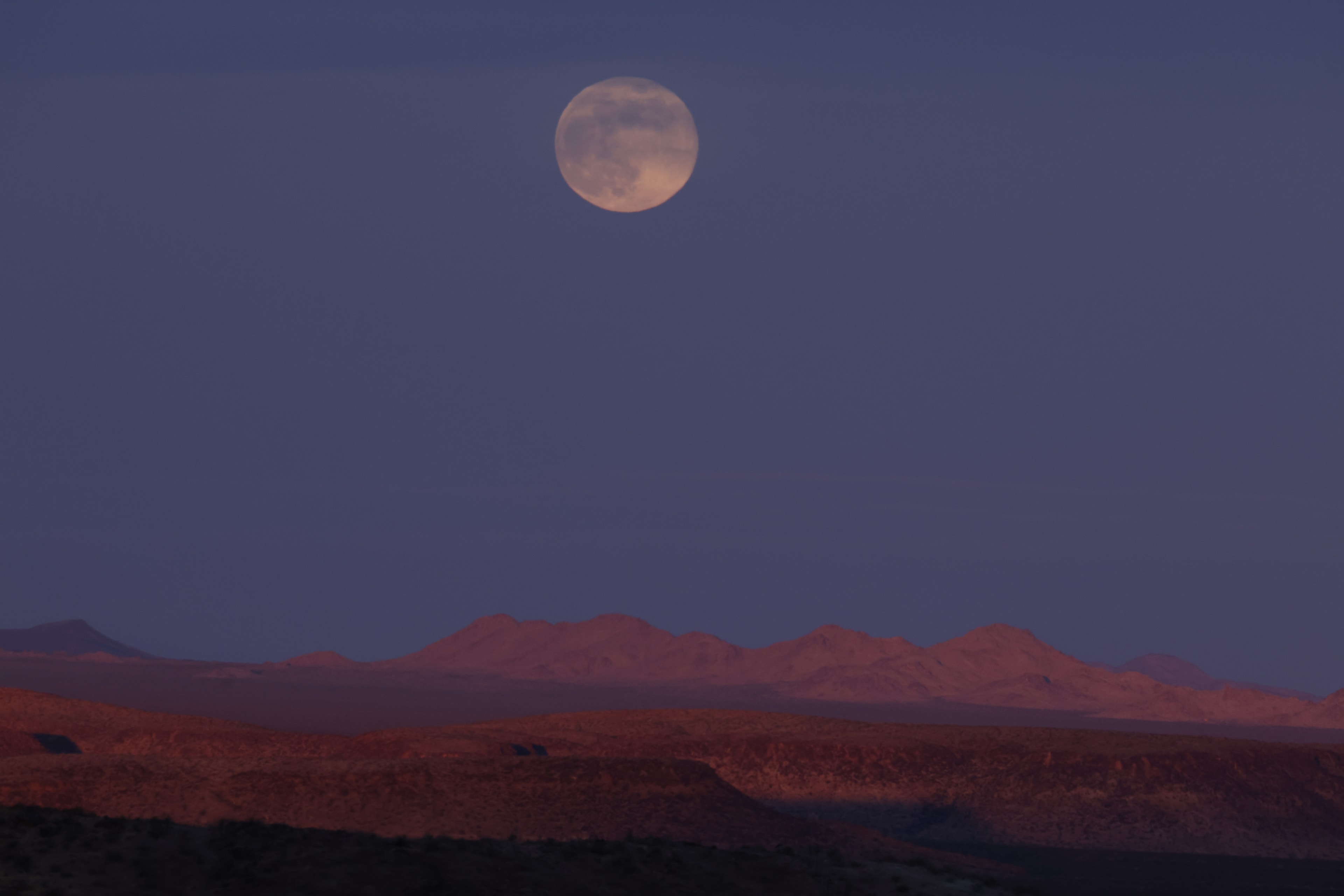 Full moon rising over desert mountains at dusk with warm red terrain
