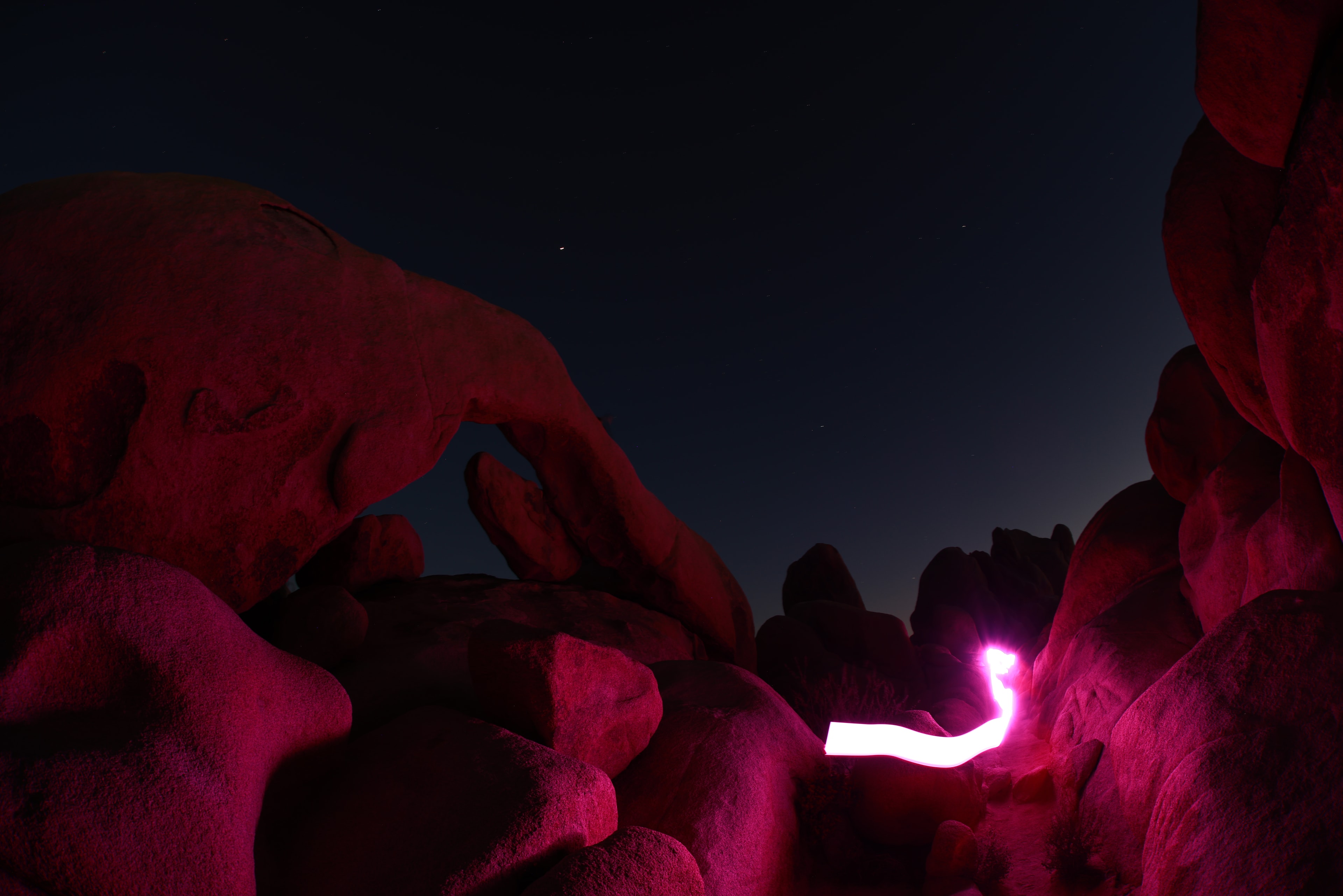 Arch rock formation bathed in magenta light with glowing light trail at night