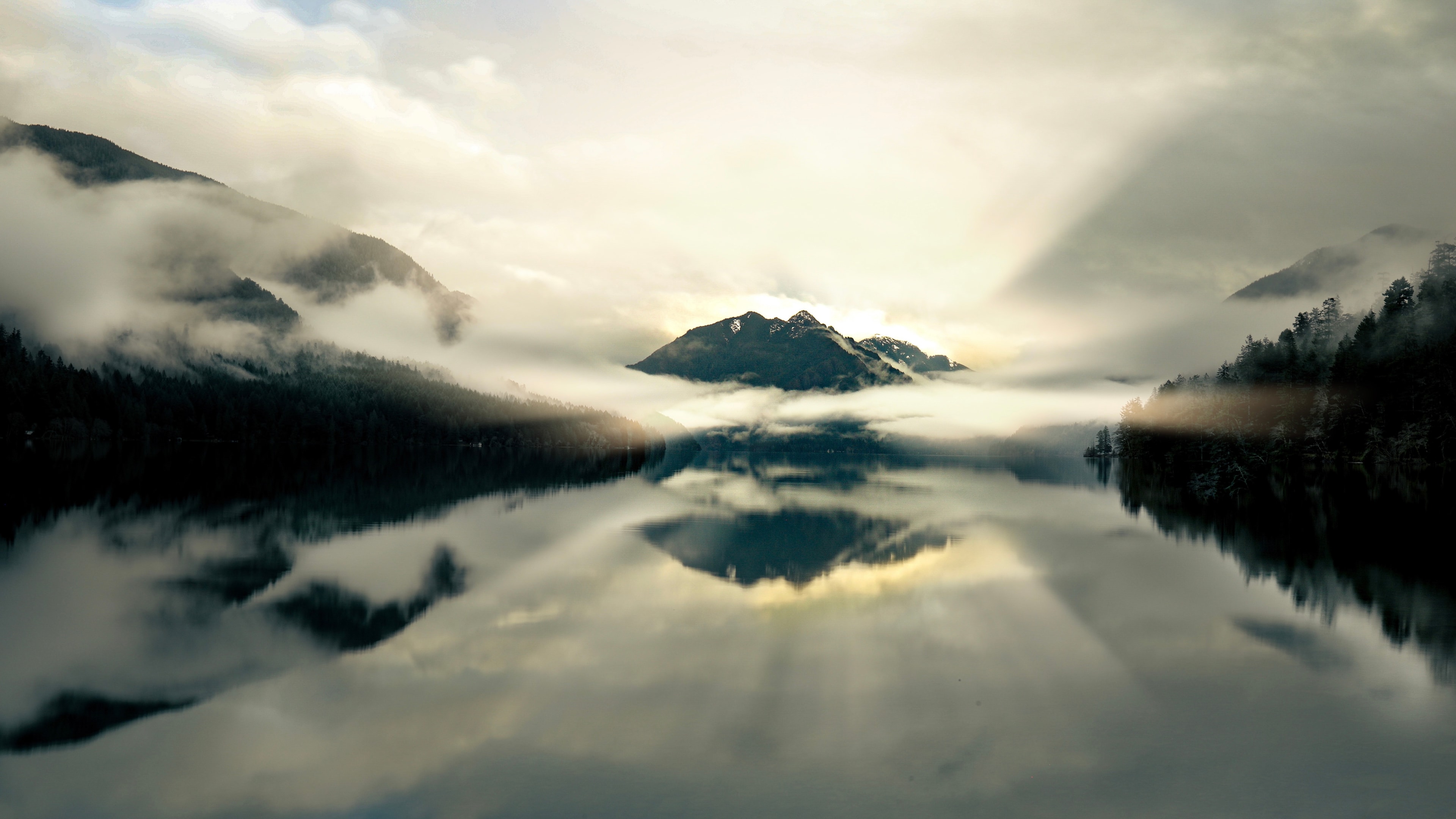 Misty mountain peak perfectly mirrored in a glassy lake surrounded by fog and forest at dawn