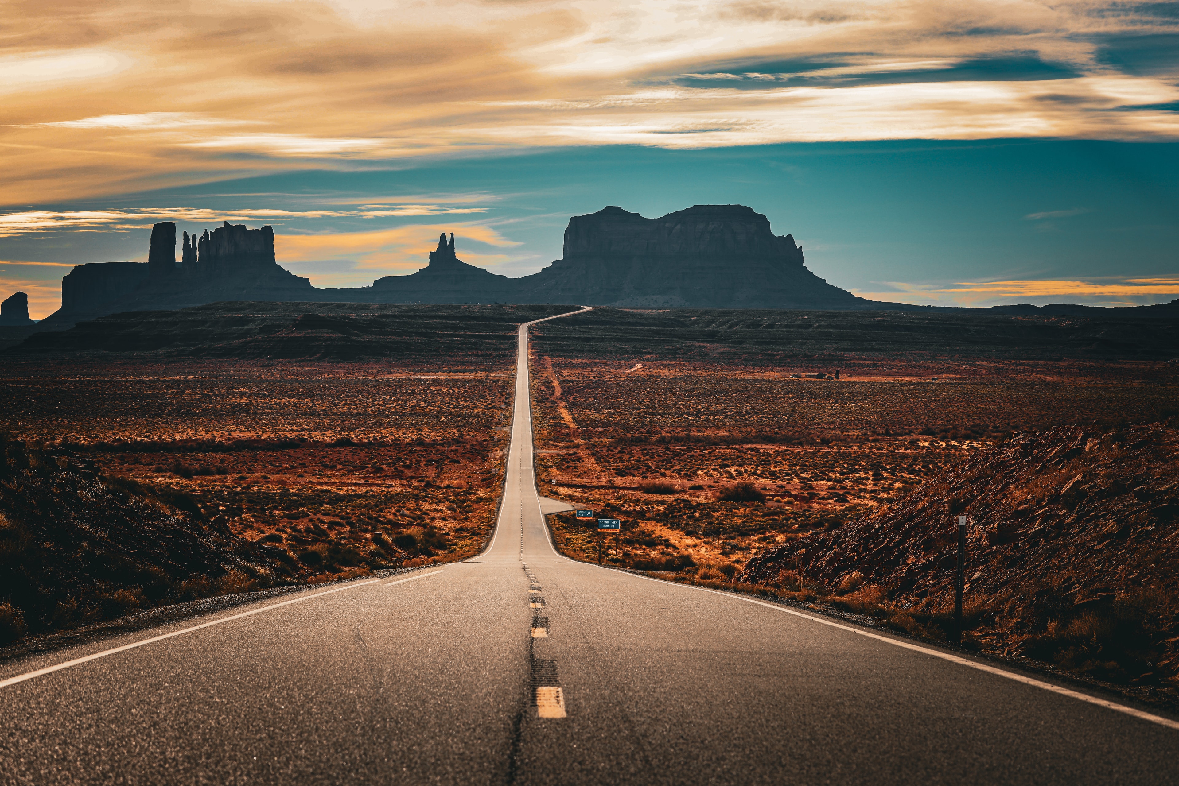 Straight desert highway vanishing toward Monument Valley buttes under dramatic sunset sky