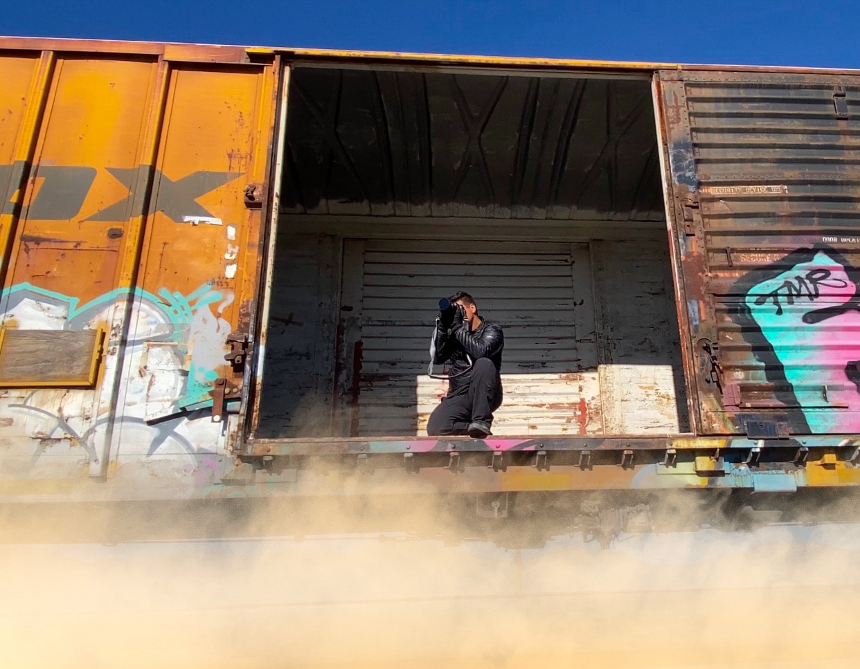 Photographer crouching inside open graffiti freight train car speeding through desert
