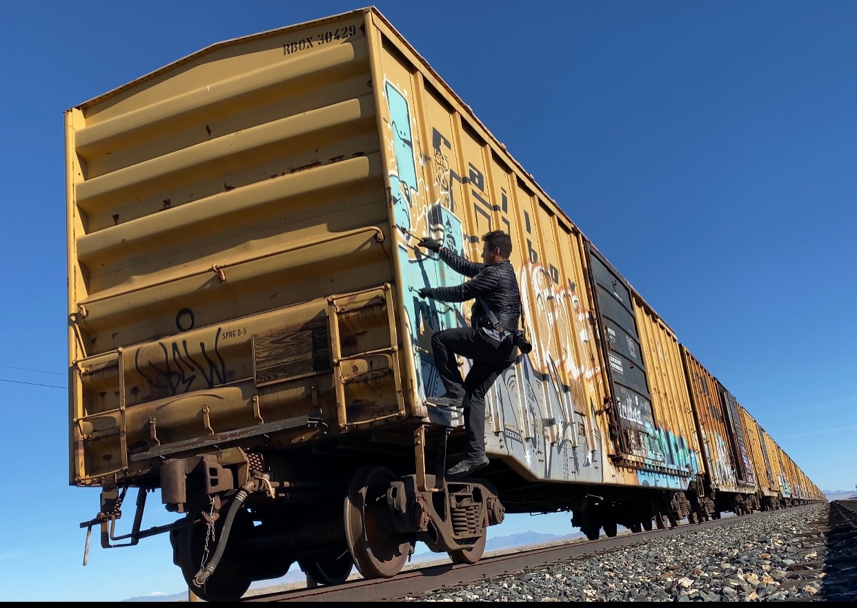 Person climbing the side of a graffiti-covered yellow freight train car under blue desert sky