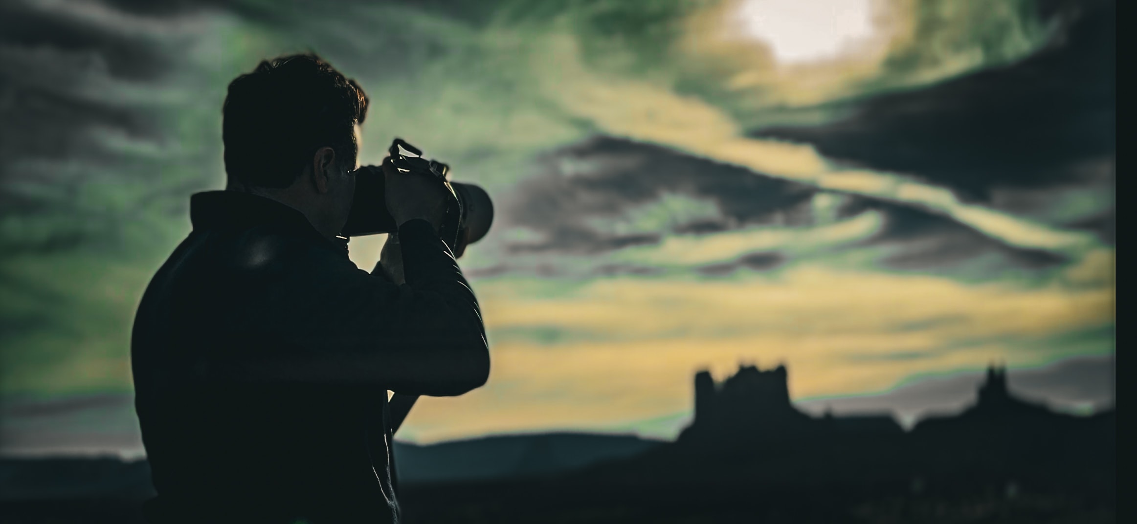 Photographer silhouetted against dramatic green and gold stormy sky with castle ruins in background
